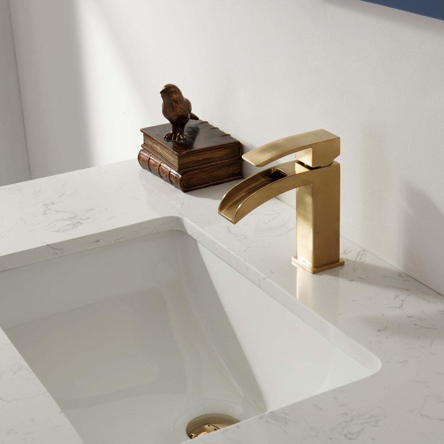 Close-up of a stone countertop with a white undermount ceramic sink and a single-handle brushed brass waterfall faucet, styled with stacked bronze books and a small bird figurine on a modern bathroom vanity.