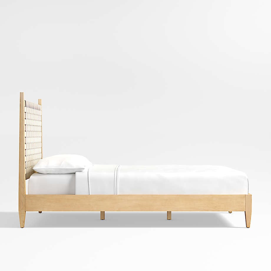 Side view of a blonde teak platform bed with a tall woven rattan headboard and low solid-wood frame, styled with crisp white sheets and a pillow in a minimalist mid-century modern design.