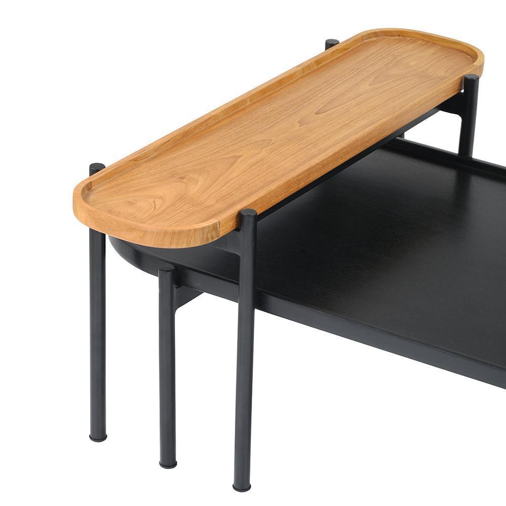 Close-up of a modern coffee table showing a natural teak oval tray top seated on a black metal frame with cylindrical legs and a lower black wood shelf.