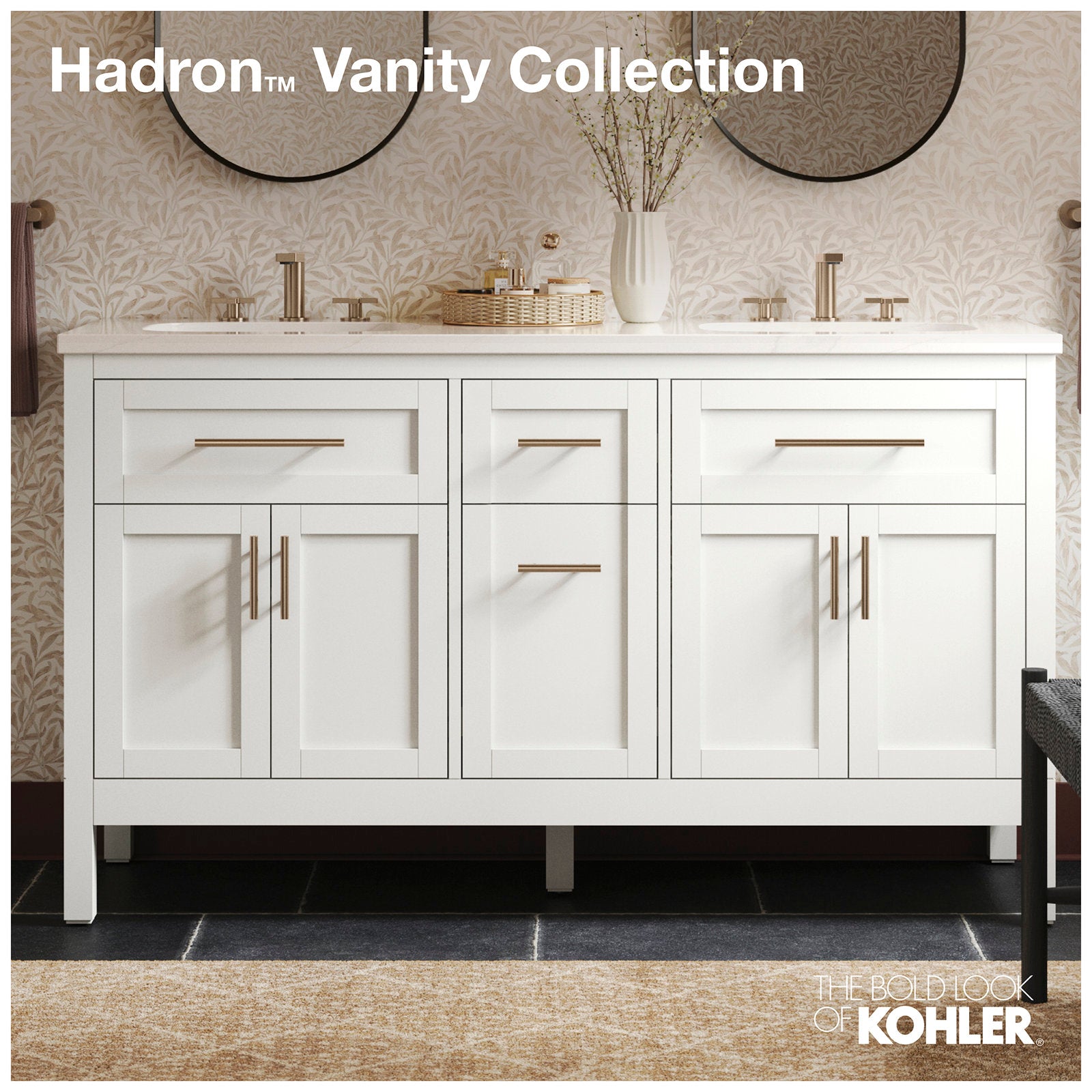 Freestanding solid wood bathroom vanity in stone finish with a stone countertop, shaker-style doors, brass bar handles, two brushed-brass faucets, round mirrors, and a vase and tray on top.