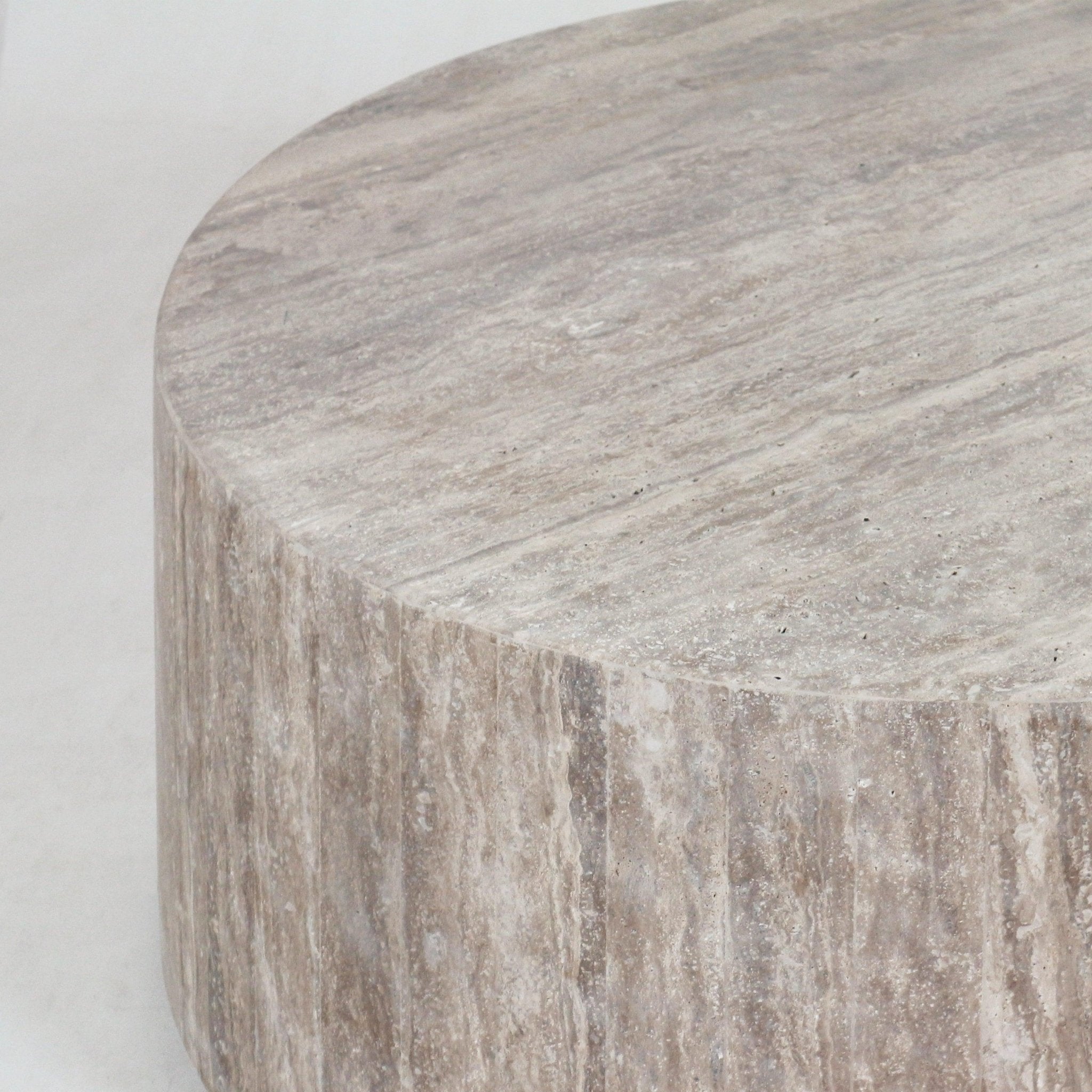 Close-up of a round travertine coffee table top and fluted side in natural beige stone, showing subtle horizontal veining and a textured contemporary finish.