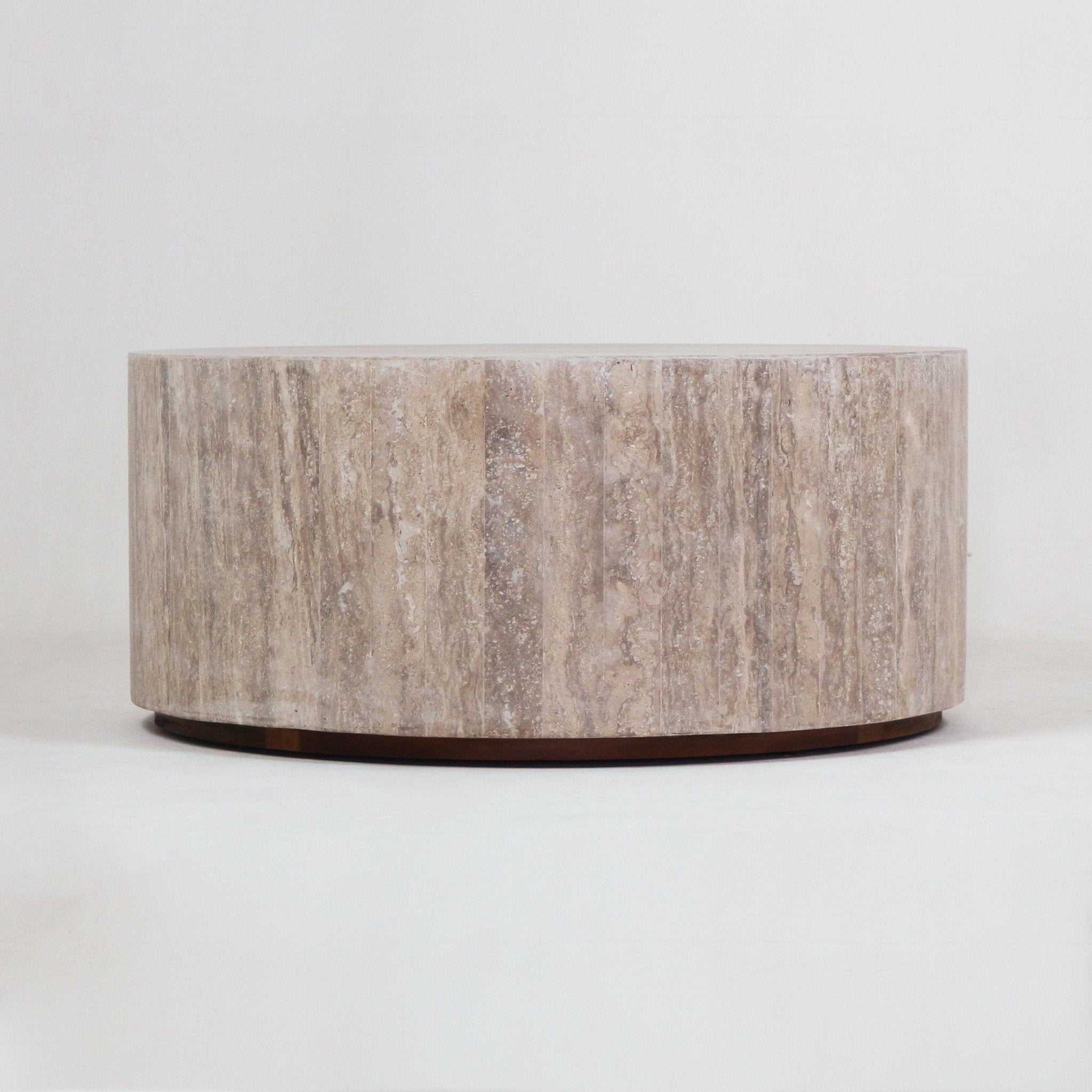 Round, low-profile travertine coffee table in natural beige with subtle vertical veining and a thin dark wood base, photographed on a white background.