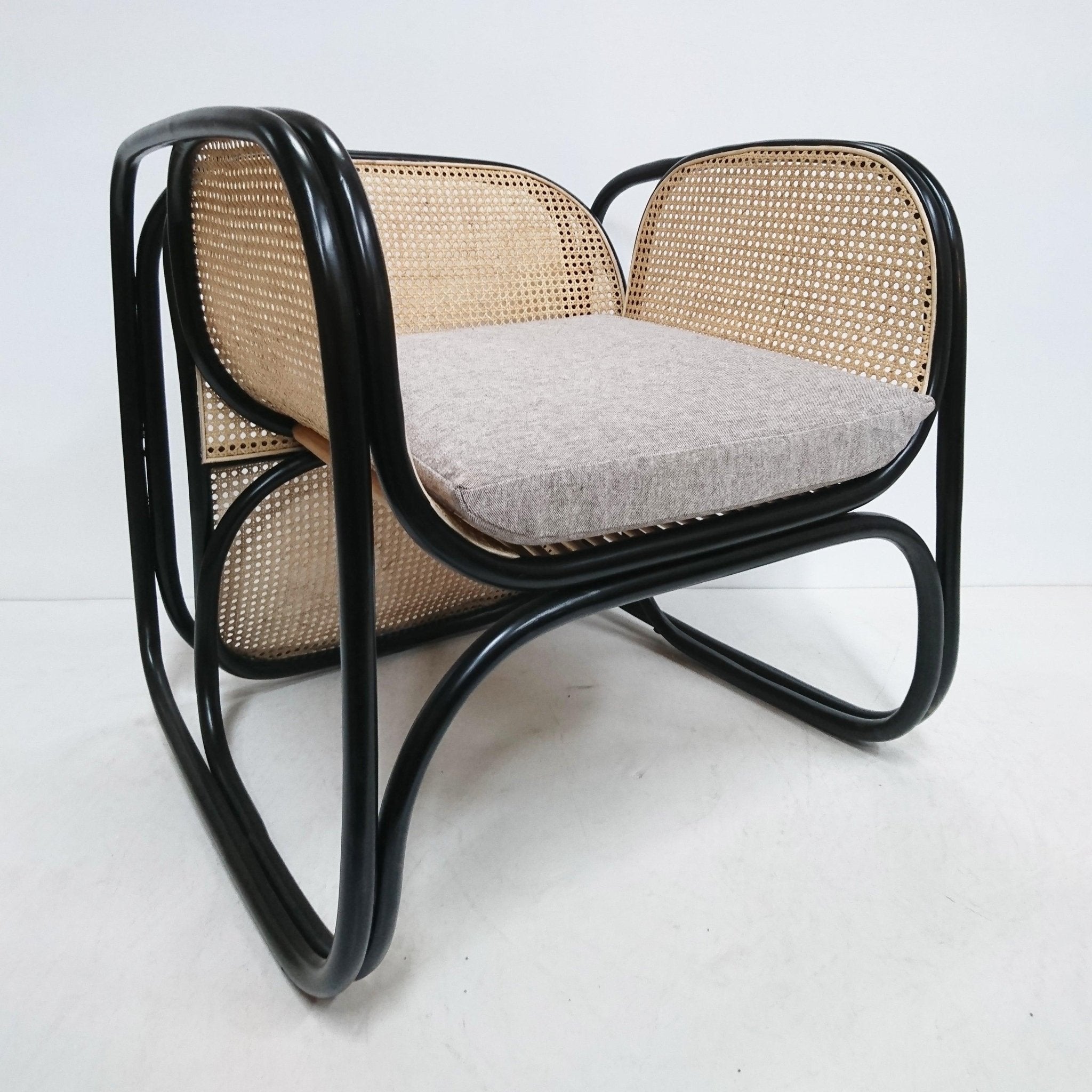 Modern cane lounge chair with a black curved tubular frame, natural woven cane back and sides, and a cushioned light gray fabric seat photographed on a white studio background.