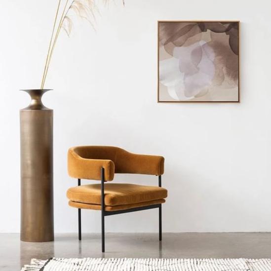 Mustard velvet modernist lounge chair with curved arms and slim black metal frame, upholstered seat cushion on concrete floor, styled beside a tall bronze vase with pampas grass and abstract wall art.