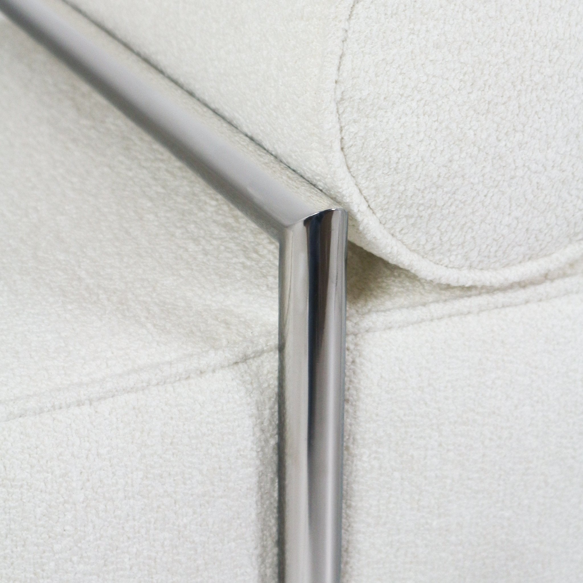 Close-up of a white boucle upholstered bench showing a round bolster cushion and polished stainless steel tubular leg and frame detail.
