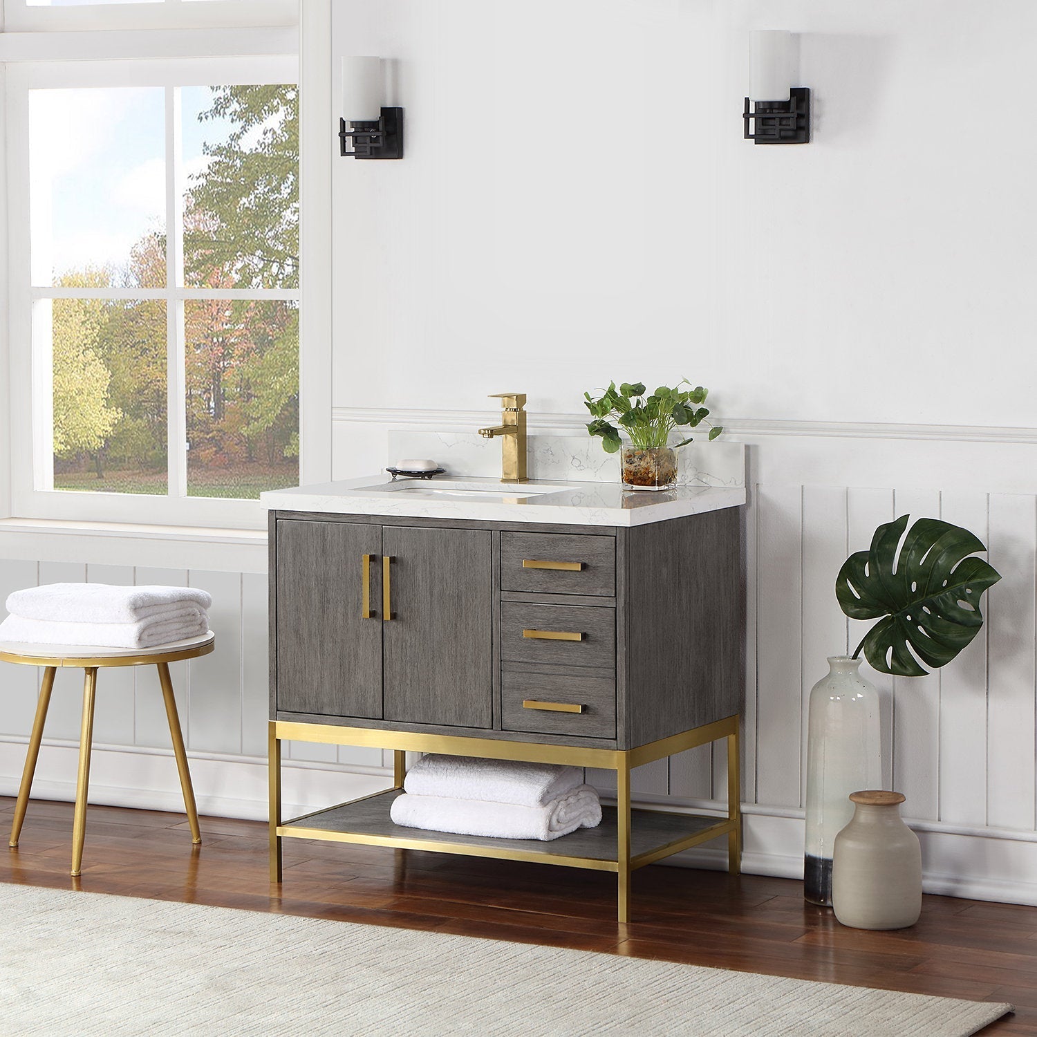Modern gray solid-wood bathroom vanity with a stone white top, single white ceramic sink and brass faucet, brass handles and metal base, three drawers, cabinet doors and open lower shelf with folded towels.