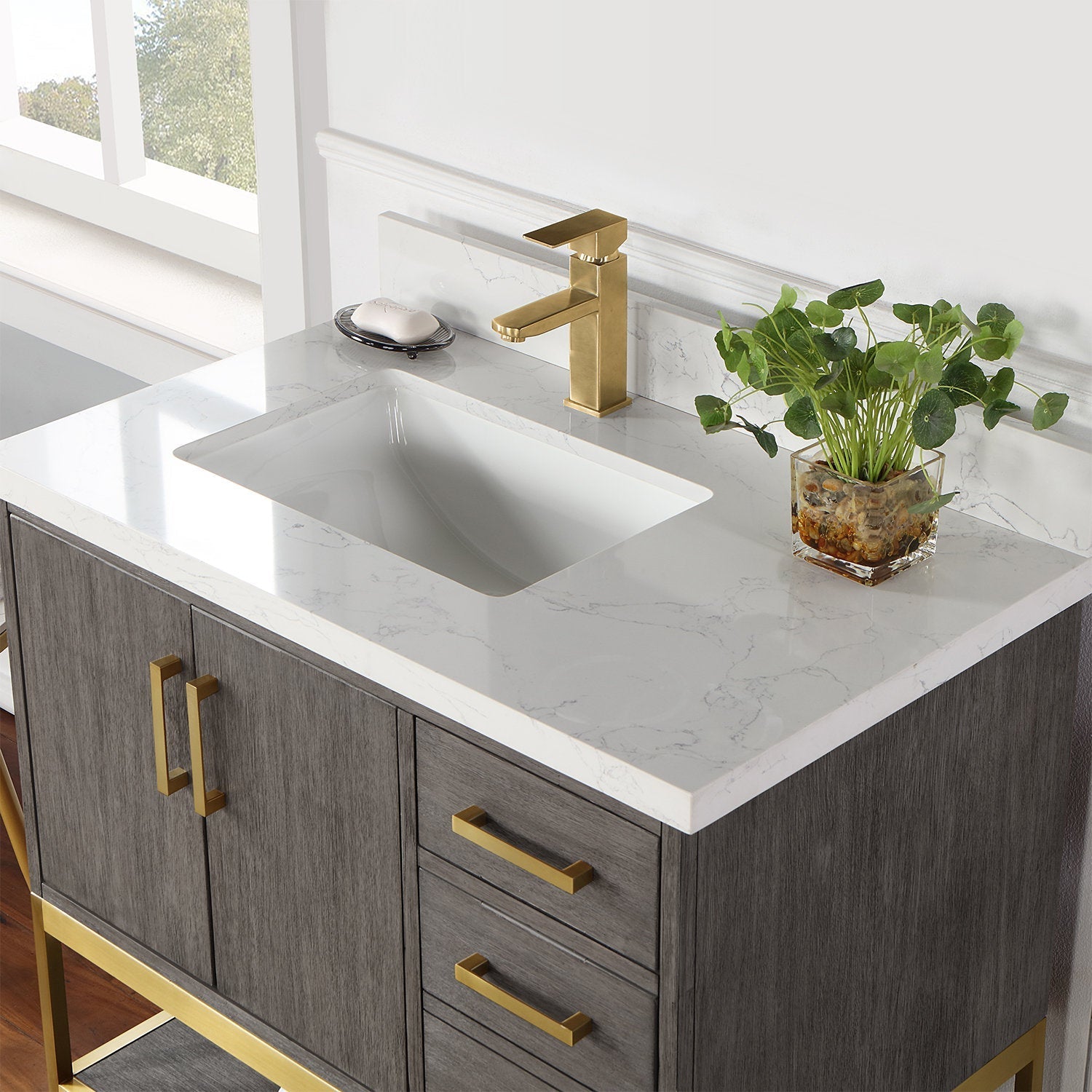 Modern 36-inch single-sink bathroom vanity with a stone countertop, undermount white ceramic sink, gold-brass single-handle faucet and matching brass handles on a gray solid wood cabinet, styled with a glass planter and soap dish.