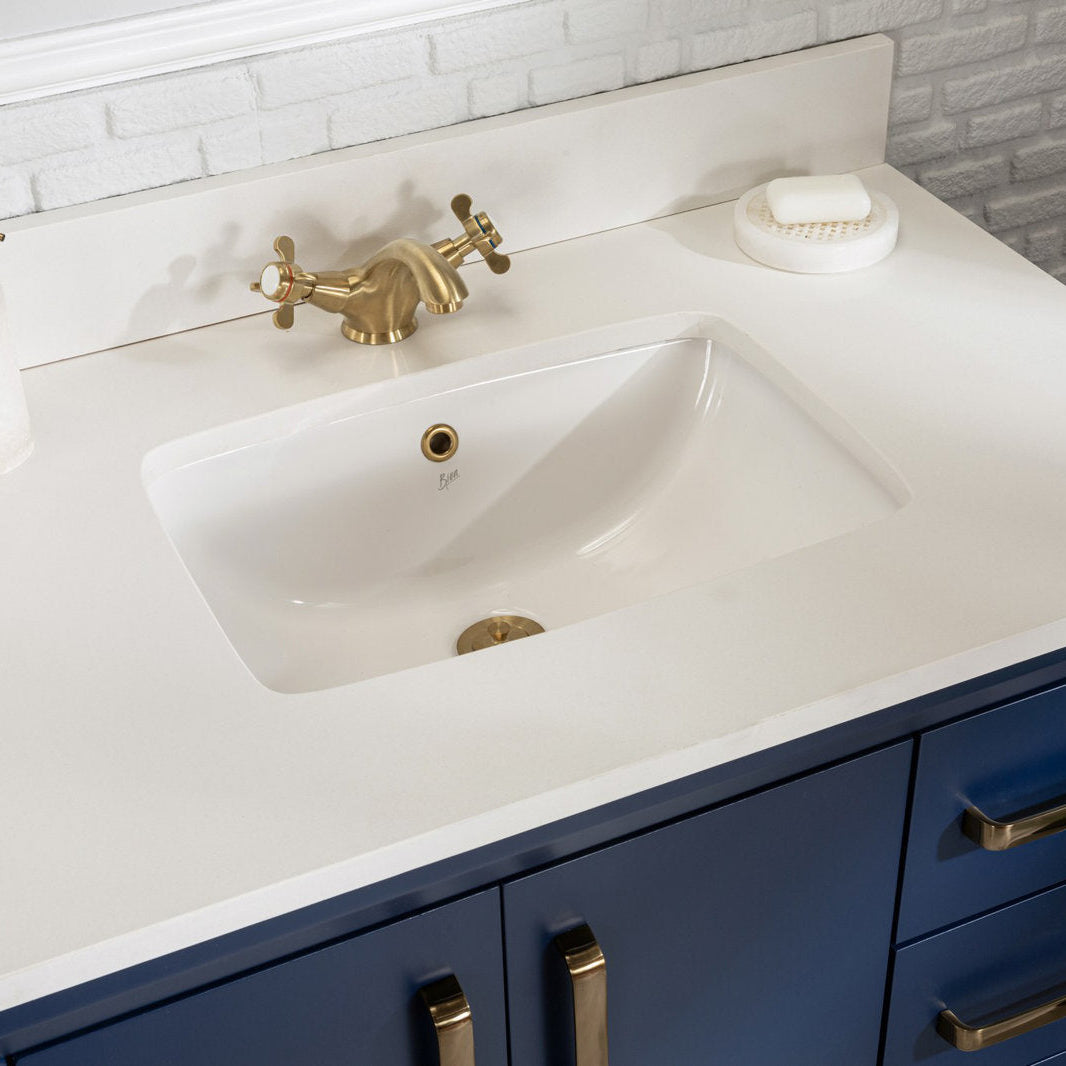 Top-down view of a white stone countertop with an integrated rectangular ceramic sink, brass cross-handle faucet and drain, navy solid-wood vanity cabinets with brass pulls and a soap dish by a white brick backsplash.