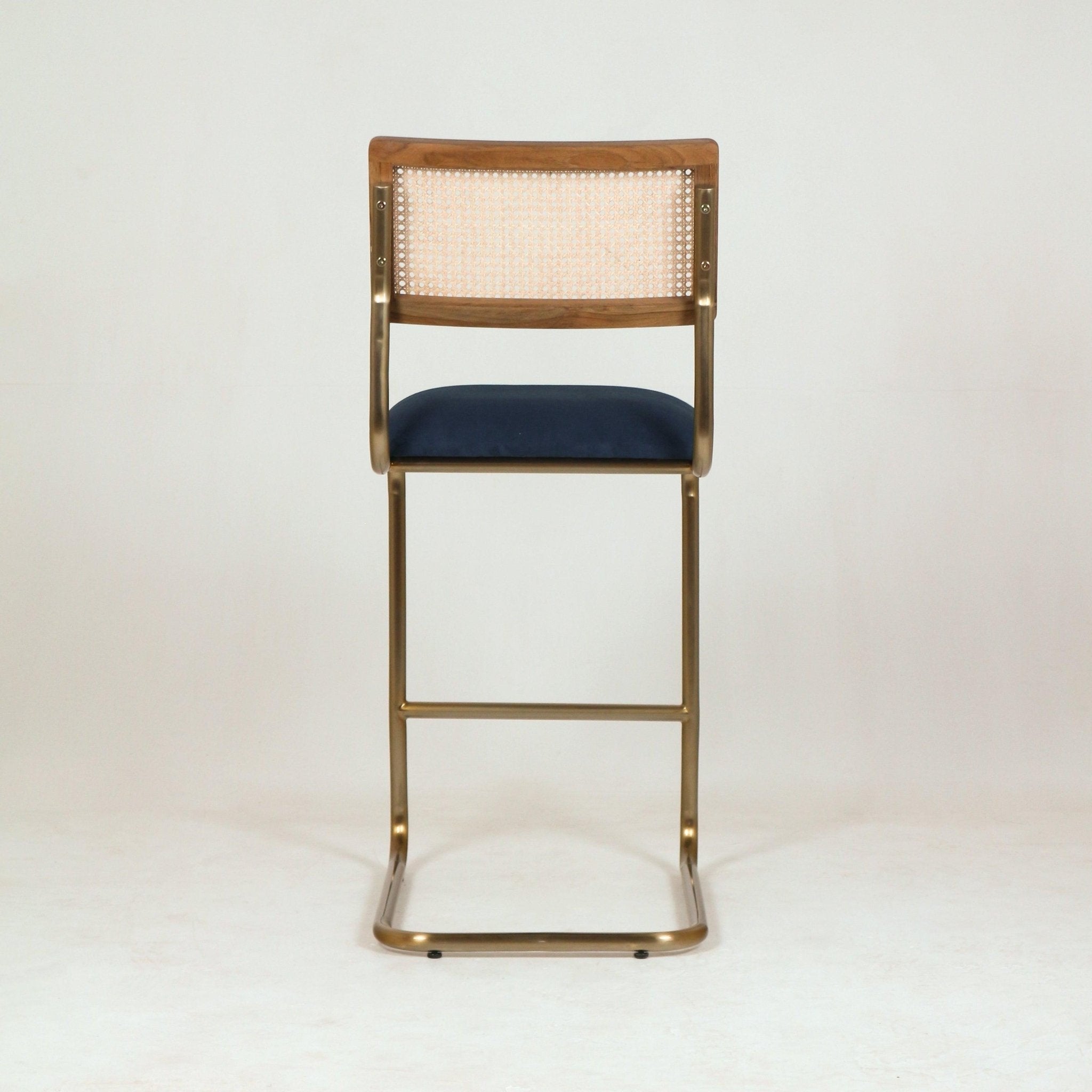 Rear view of a modern cantilever bar stool with a brass metal frame, navy blue upholstered fabric seat, natural cane back panel and walnut wood top rail against a neutral studio background.