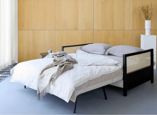 Modern sofa bed with a black wooden frame, cane rattan panels and slender metal legs, dressed in neutral beige linens and gray pillows, shown against a light wood-paneled wall with bedside decor.