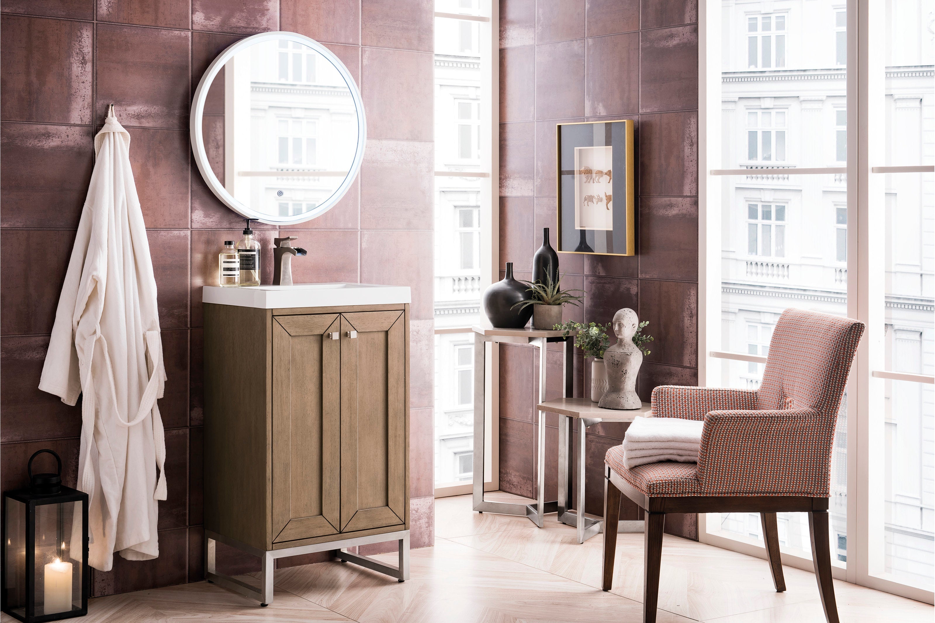 Freestanding modern solid wood single-sink bathroom vanity in a stone finish with a white stone top and metal base, shown with a round mirror, faucet, hanging robe, lantern, side tables and upholstered chair against mauve tiled walls.
