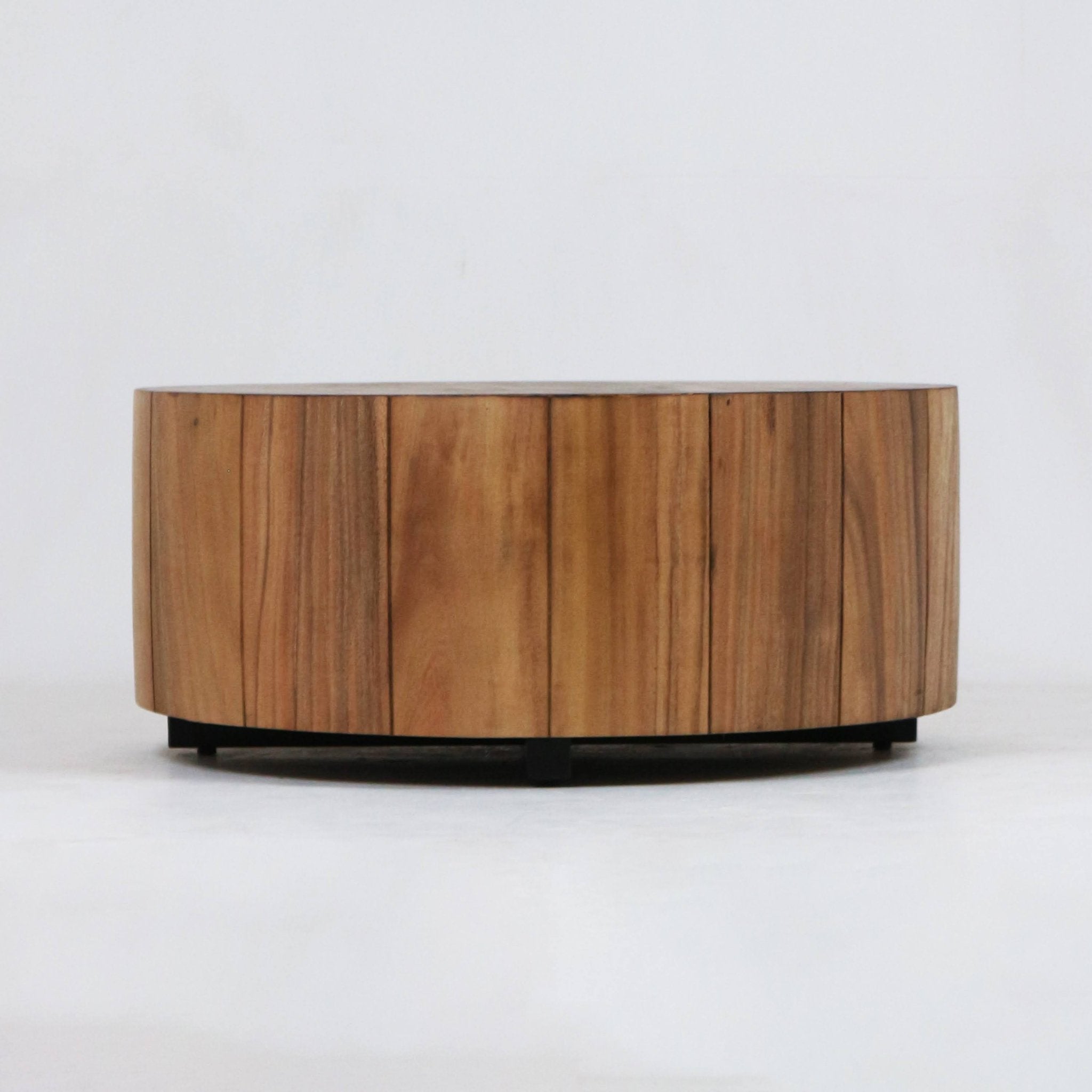 Low round solid wood coffee table with vertical plank panels and visible natural wood grain, supported by a recessed black metal base in a modern minimalist style, photographed on a white background.