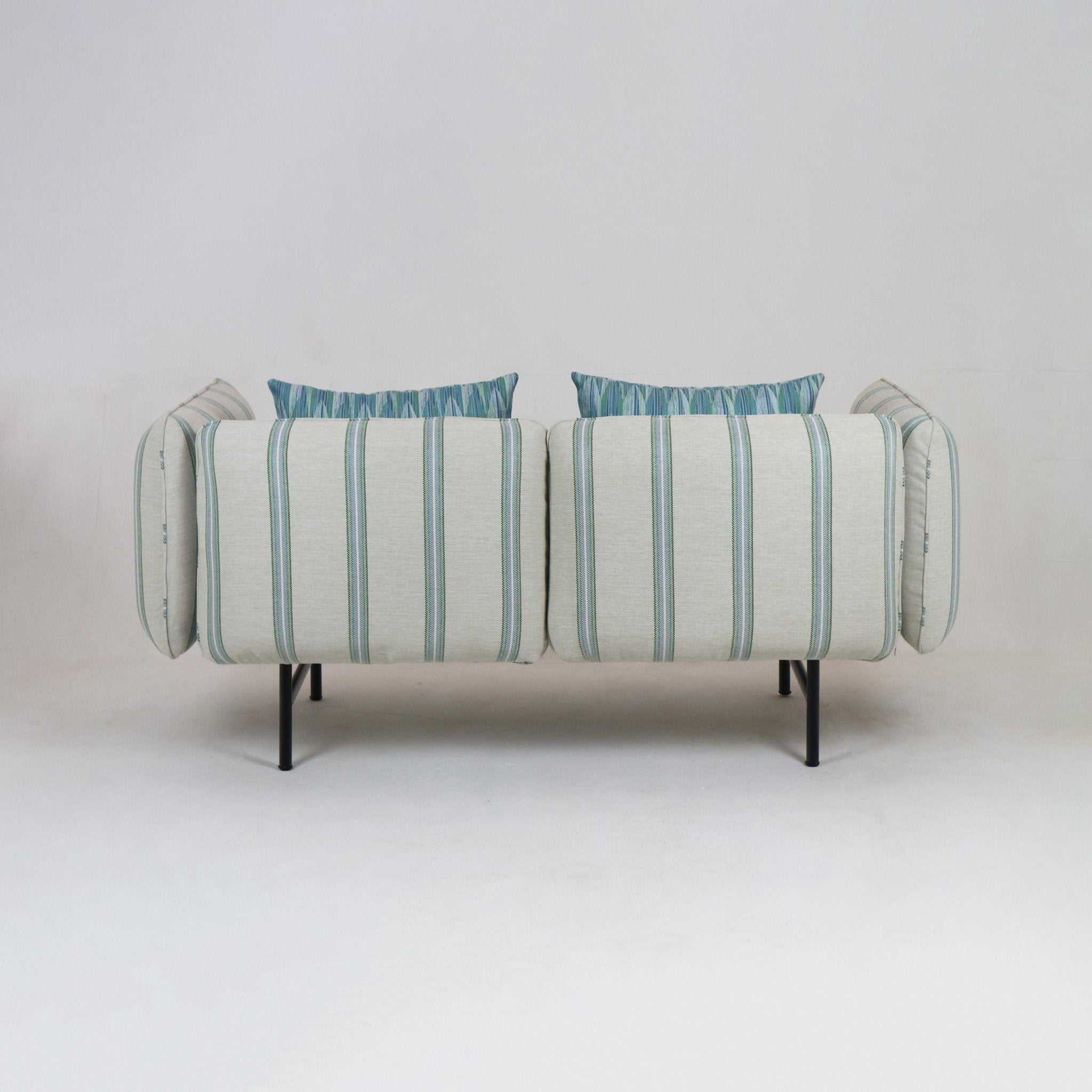 Rear view of a coastal outdoor sectional sofa upholstered in light striped fabric with green and blue accents, blue-green patterned back cushions, teak-finish frame and slim black metal legs.