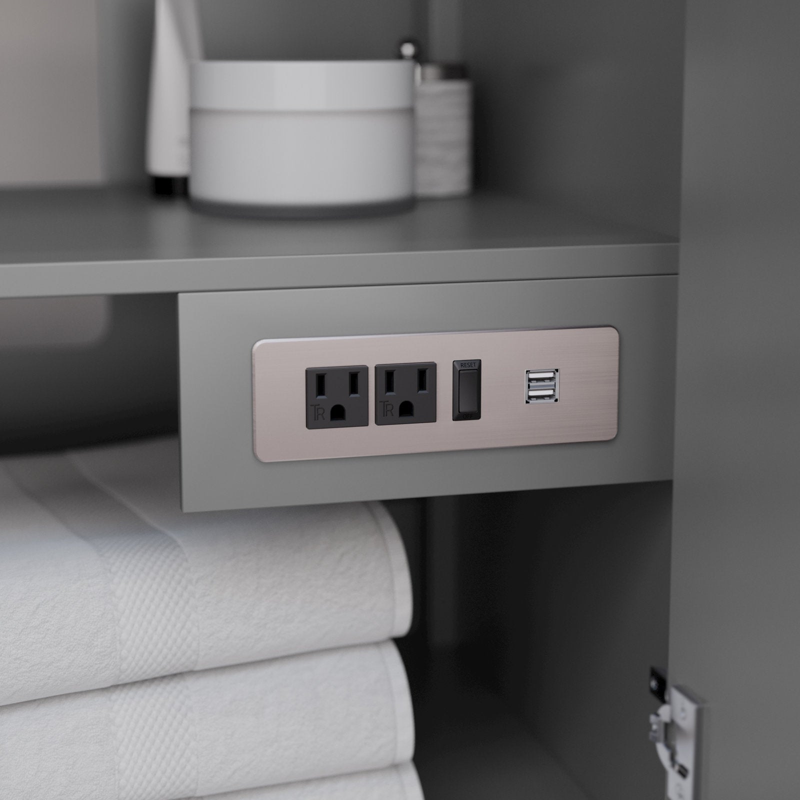 Built-in brushed-metal outlet panel inside a stone-colored modern vanity cabinet showing two standard electrical sockets, a reset/GFCI switch and dual USB ports with folded white towels on the lower shelf.